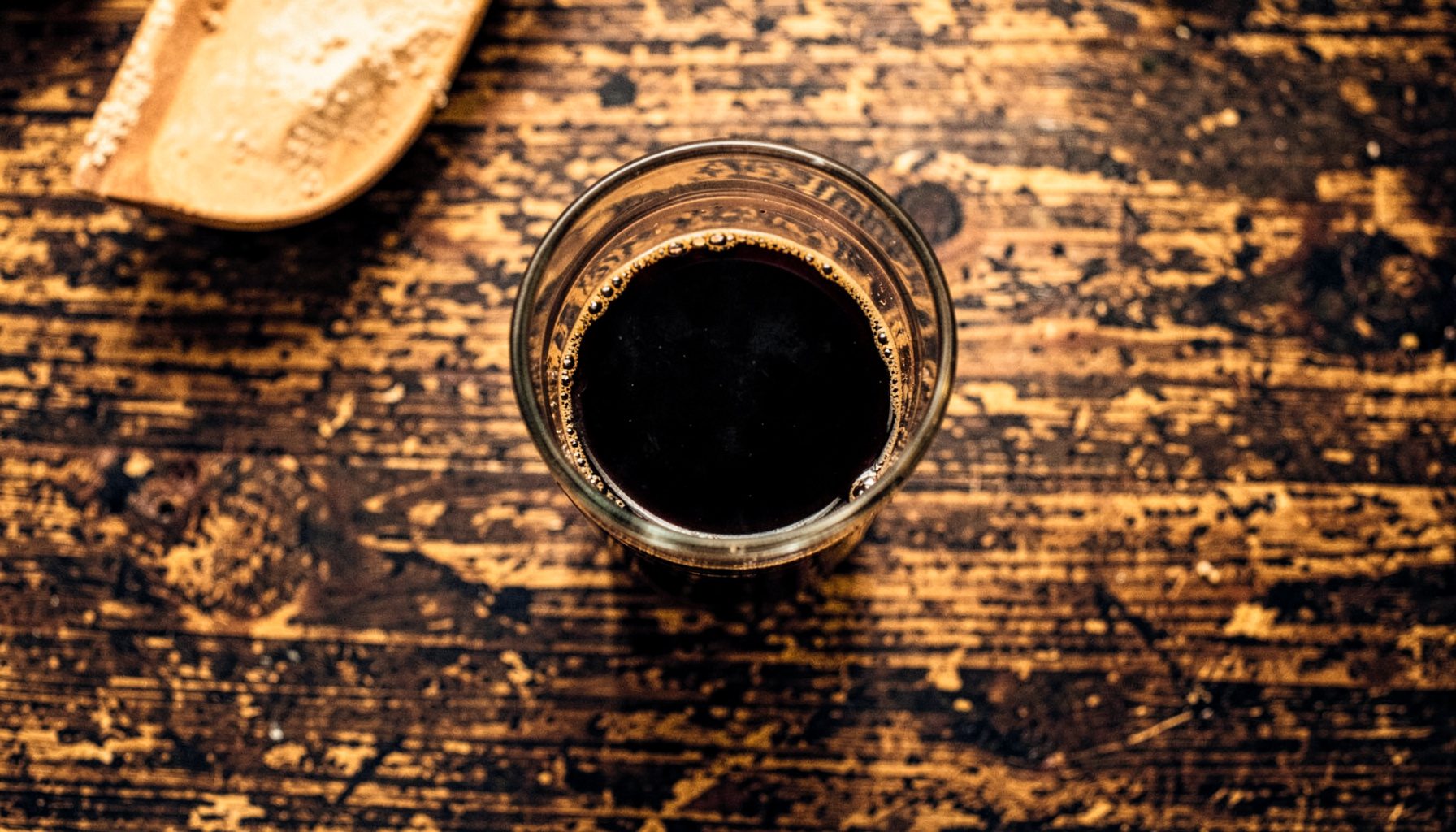 Overhead flat-lay of a dark stout in a glass on a worn wooden pub table, deep black and cream tones with warm amber light, cozy and unhurried mood